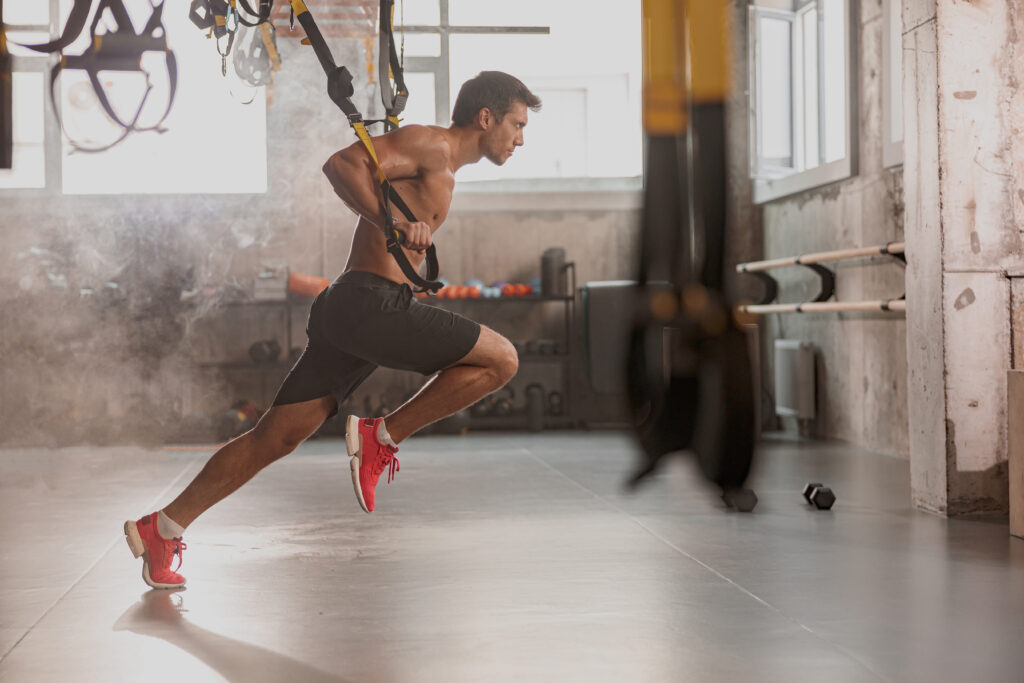 Side view of athlete sporty man doing exercise with fitness straps to strengthen his muscles of hands in gym